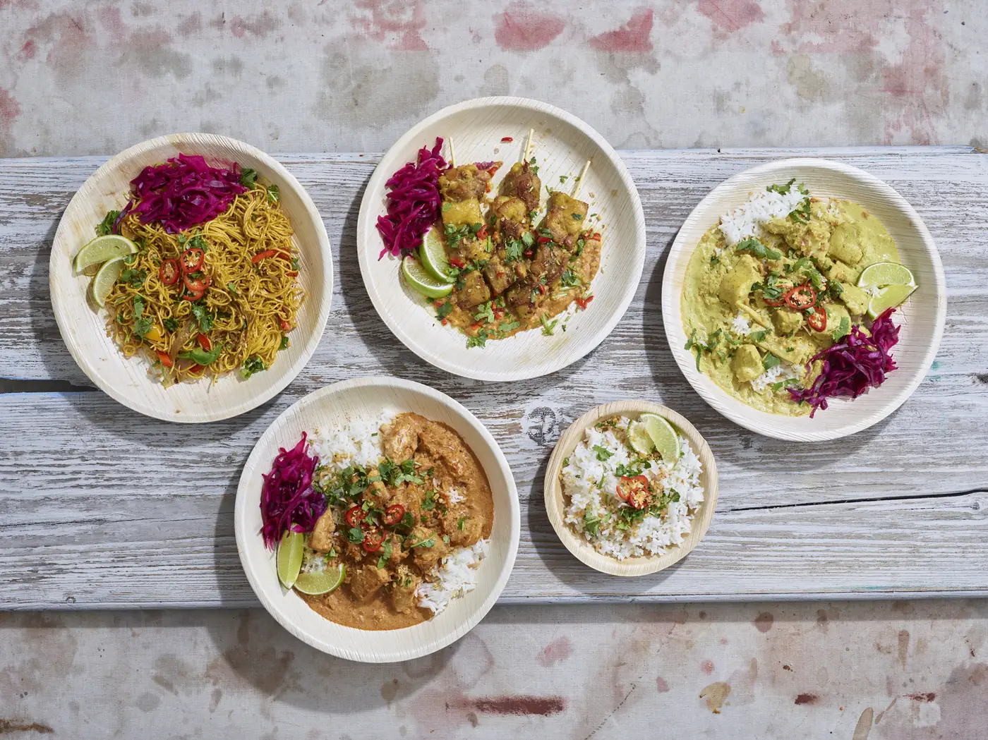 A spread of Tippy's Thai ready meals — stir-fried noodles, chicken satay skewers, yellow curry, panang curry and jasmine rice, each served with pickled red cabbage and lime